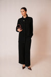 A woman modeling a black long-sleeve collared jacket and matching wide-leg trousers, holding a dark brown woven clutch against a plain beige studio background.