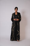 A woman modeling a long black abaya with a gold marble vein pattern and a matching open-front jacket, standing against a plain light gray studio background.