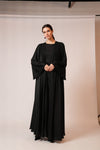 A woman modeling a long black abaya dress with wide draped sleeves in a studio setting with a light beige backdrop.